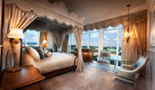 A 4 poster king bed with a canopy, a night stand, and a glass table with 2 padded arm chairs
A 4 poster king bed with a canopy, a night stand, and a glass table with 2 padded arm chairs