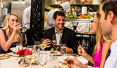 Two men and 2 women enjoy dinner as chefs prepare food in an open kitchen
Two men and 2 women enjoy dinner as chefs prepare food in an open kitchen