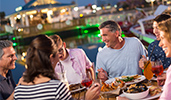 Six adults enjoy dinners and appetizers at an outdoor table next to a lake
Six adults enjoy dinners and appetizers at an outdoor table next to a lake