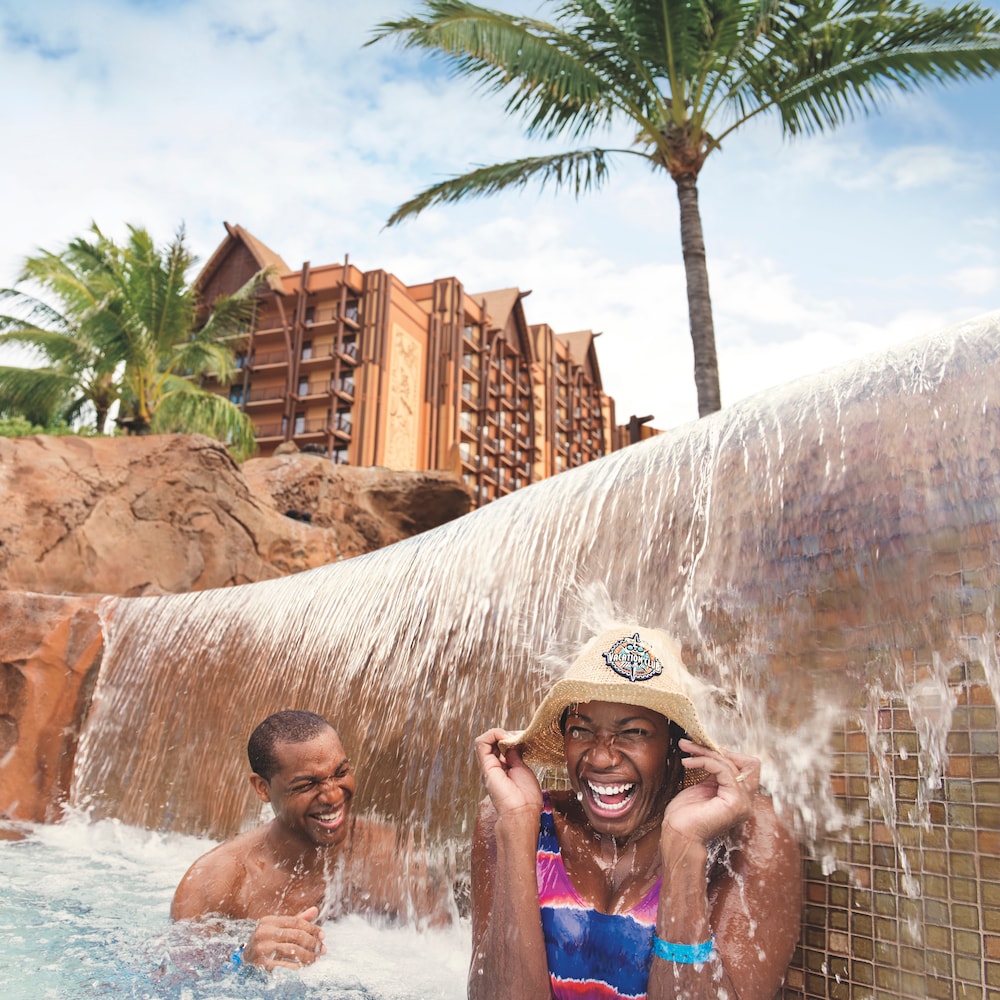 A man and a woman holding onto her hat are all smiles as they stand under a waterfall at an Aulani Resort pool