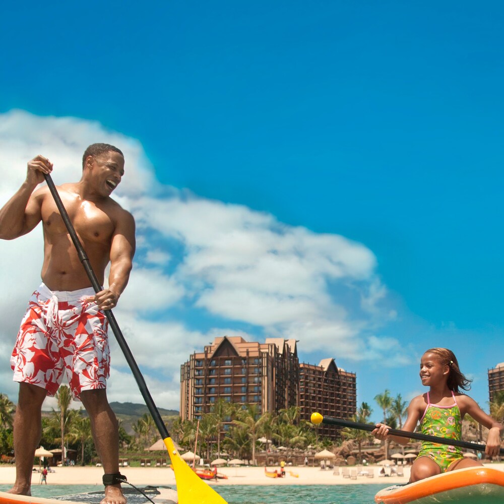 A father stands on a paddleboard next to his daughter, who is kneeling on a paddleboard, on the ocean in front of Aulani Resort