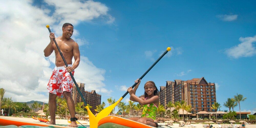 A father stands on a paddleboard next to his daughter, who is kneeling on a paddleboard, on the ocean in front of Aulani Resort