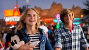 A young couple, with their stuffed Mickey doll, walk along the neon-lit street of Radiator Springs in Cars Land at nighttime