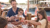A young girl cracks a lobster claw, as her brother and father sit with her at a dockside table containing a beer and a plate of lobster