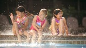 Three smiling young girls sit at the edge of a swimming pool and splash water around