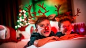 Two boys smiling on their hotel room bed each wearing antlers and a red nose