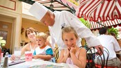 A Carnation Cafe chef visits a mother and 2 children as they dine at this outdoor Disneyland restaurant