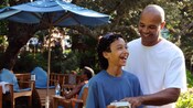A father and son laugh together as they view a photograph on a camera in front of a shade umbrella and grouping of chairs