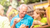 A young boy and his grandparents enjoy pulled pork sandwiches at an outdoor table