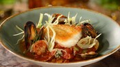 A bowl containing Cioppino seafood stew includes grilled bass, mussels, clams and herbs in a tomato based broth