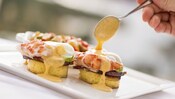A hand drizzling Hollandaise sauce from a spoon over lobster eggs benedict