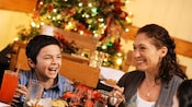 Mother and son smiling and laughing at a holiday feast, with a Christmas tree behind them