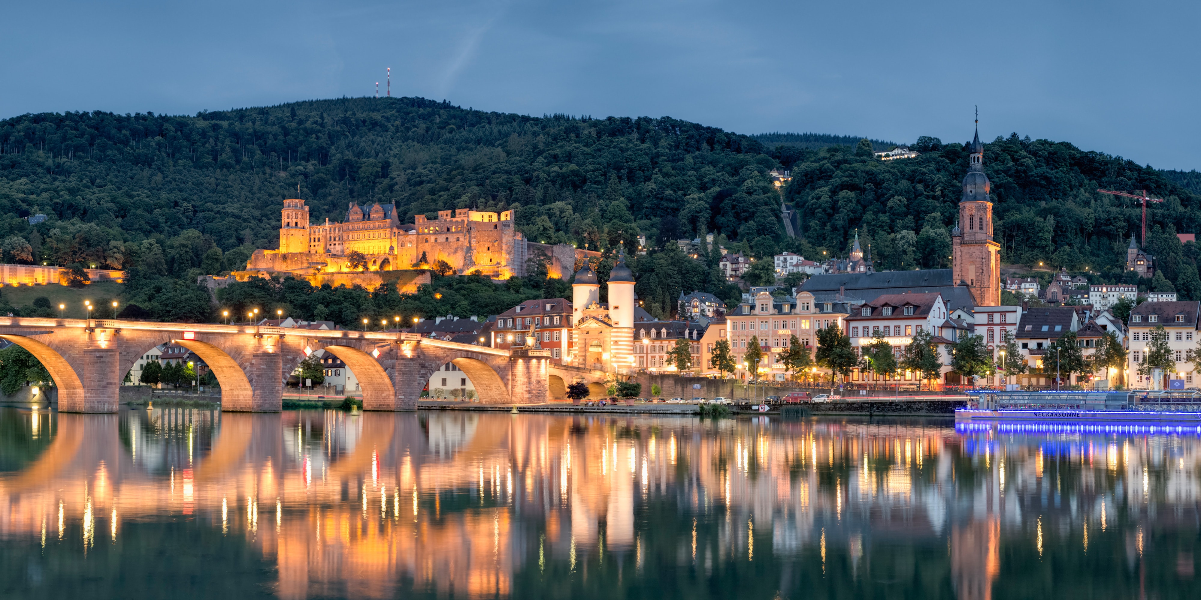 Heildelberg Castle stands between a tree-lined mountain and and a stone bridge, which is reflected in the Rhine River
