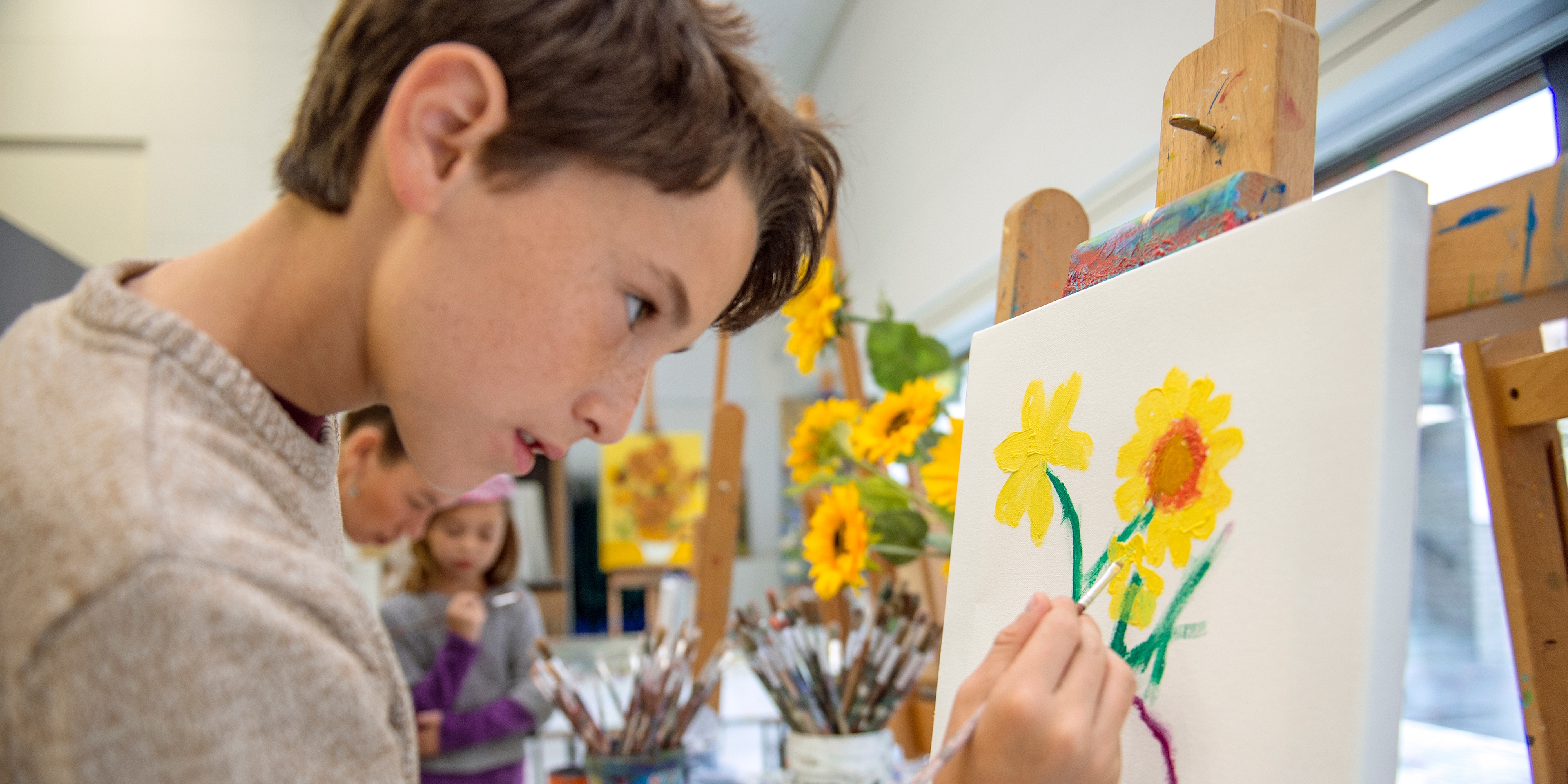 A boy stands at an easel creating a painting of sunflowers, copying the sunflowers in a nearby vase