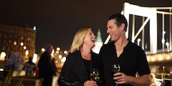 A man and a woman stand on a ship's deck at night, drinking wine and laughing