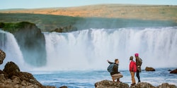 3 people stand on the rocks admiring Goðafoss Waterfall