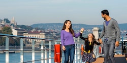 Parents and their young daughter laugh while walking on the ship’s deck as they sail past Budapest