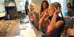 A girl with earphones sits in front of a microphone at a radio control booth, with others watching