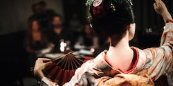 The back of a Japanese maiko in traditional dress, who is dancing with a fan in front of an audience