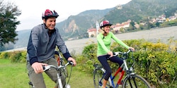 A pair of Guests ride bikes along a riverbank in front of rolling hills and historic buildings