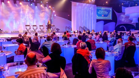 A woman speaks on a stage while an audience seated at tables with glasses of wine and small plates of food listens. Over the stage, a sign reads The Making of Party for the Senses, from idea to implementation