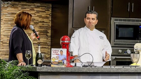A woman with a microphone speaking to Chef Buddy Valastro in a demonstration kitchen containing a mixing machine and one of the chef’s cookbooks