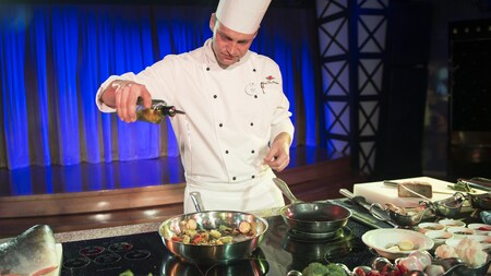 A chef in a demonstration kitchen pours oil into a sauté pan containing scallops and vegetables