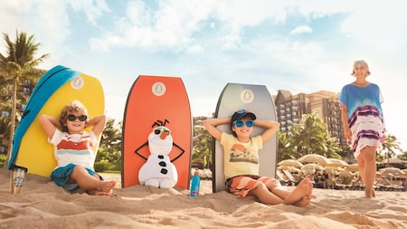 Two boys sit on the beach with Olaf the snowman, while their grandmother stands nearby
