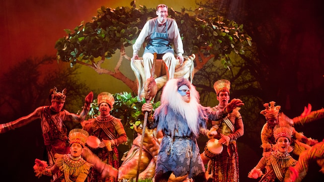 A father watches from a tree as Rafiki from The Lion King shares wisdom in a song accompanied by drums