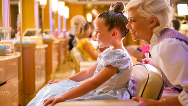 A girl admires her princess makeover with a Fairy Godmother in training, at Bibbidi Bobbidi Boutique