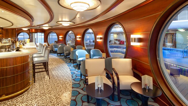 A lounge has high stools at a casual bar and rows of chairs with cocktail tables next to porthole windows