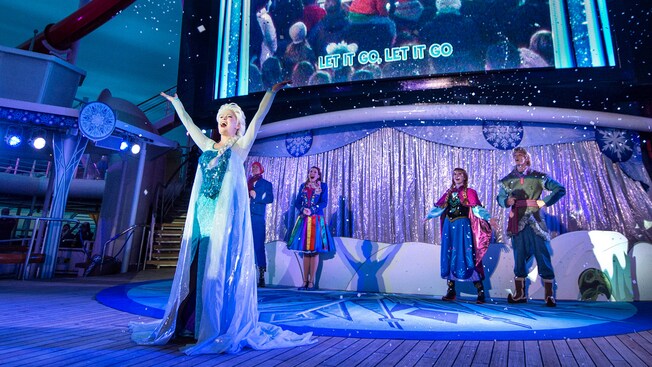 The lyrics to Let It Go appear on a screen behind Elsa as she leads a crowd of Guests in a singalong