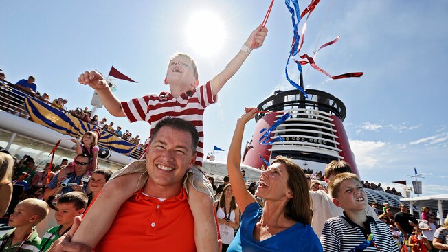 A family standing amid a crowd gathered on the deck of a cruise ship celebrating with streamers