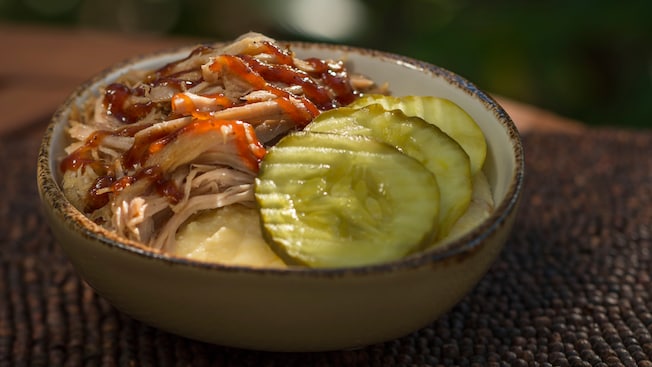 A bowl with pulled pork and pickles, with cheddar grits peeking out from the bottom