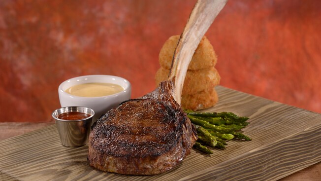 Dry-aged long bone rib chop plated on a wooden cutting board with sauces, onion rings and asparagus