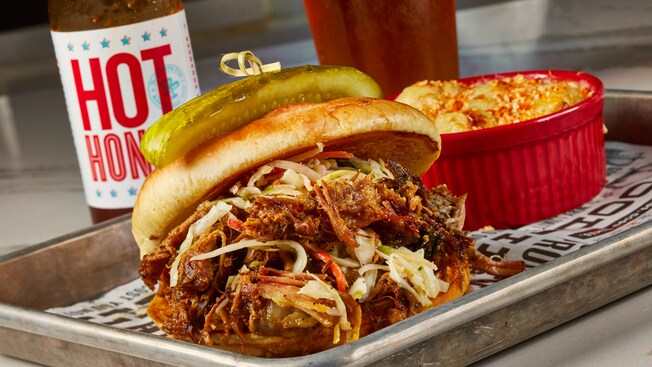 A tray with a pulled pork sandwich with a pickle on top adjacent to a mini casserole dish of mac and cheese