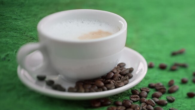 A foamy cup of cappuccino from STARBUCKS® plated beside a scattered array of espresso beans