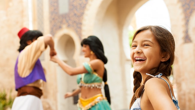 A girl in pigtails smiles while Aladdin and Jasmine play in the background at Epcot