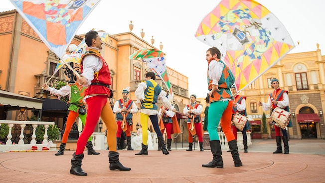 Men in colorful outfits wave multicolored flags as they are backed by live musicians