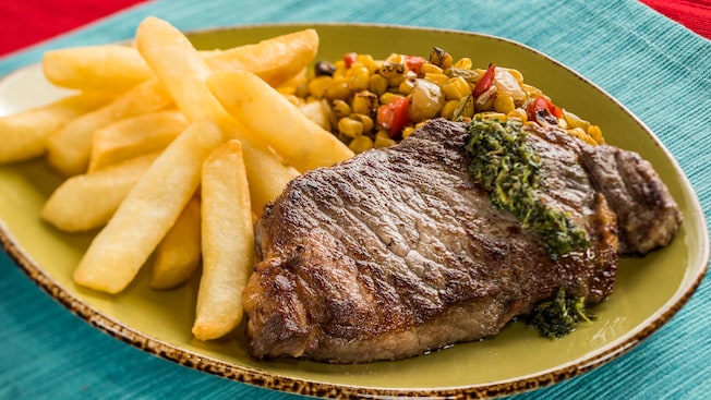 A steak topped with chimichurri sauce is served with french fries and succotash