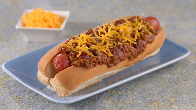 A hot dog covered in chili and shredded cheddar cheese on a plate near a dish of cheddar cheese