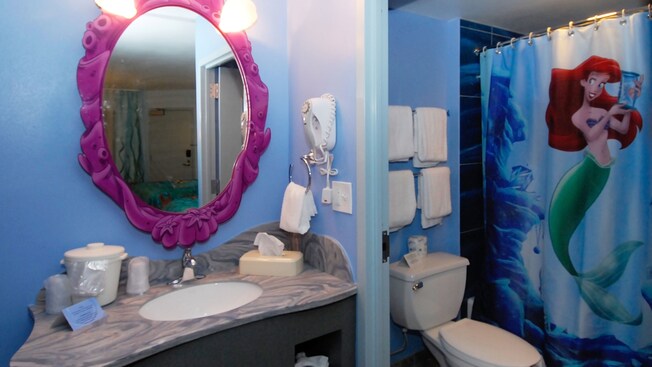 A vanity with a mirror above a countertop sink next to a toilet and a bathtub/shower with a curtain featuring Ariel from Disney's 'The Little Mermaid'