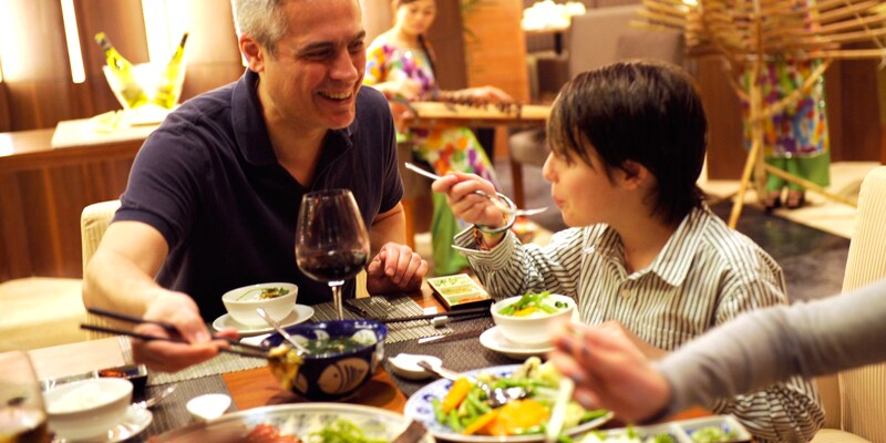 A boy blows on his hot soup to cool it as his father eats from a bowl with chopsticks