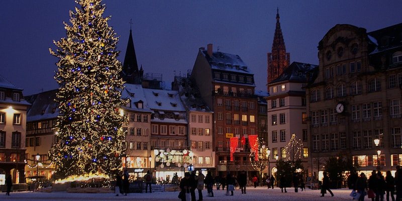 At night, lights twinkle on a tall Christmas tree in a square with pedestrians and quaint shops