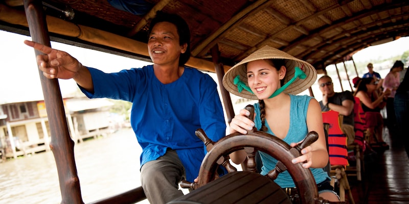The driver of a roofed touring boat cruising on a river lets a female tourist take the wheel