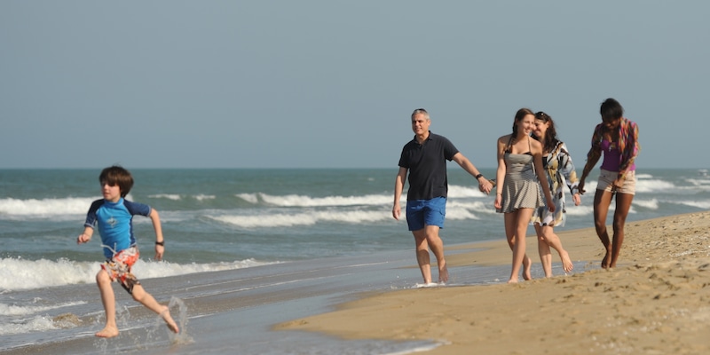 A boy splashes in the waves as he runs ahead of 2 couples walking on a beach
