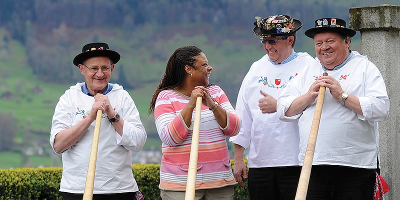 A woman stands with 3 costumed Swiss alpine horn players and laughs as she takes one of their horns