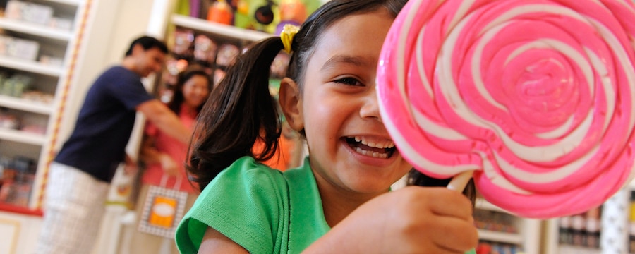 A young girl in a candy shop smiles as she holds her giant pink and white swirled lollipop and her parents shop in the background
